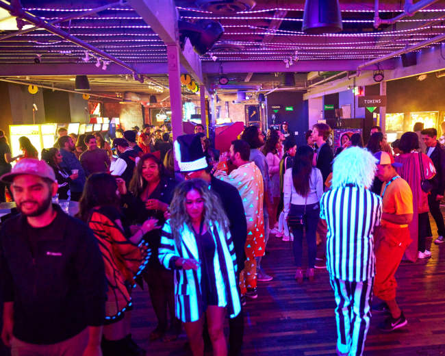 A large crowd of people in various costumes gathers in a brightly lit arcade with games and tokens visible in the background.