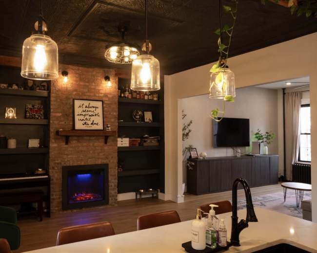 The image shows a modern living space featuring a kitchen island, an electric fireplace with a brick surround, open shelving with decorative items, and pendant lighting above the island.