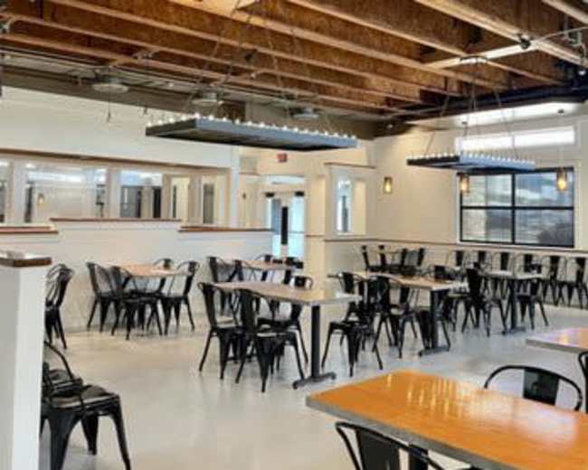 The image shows a spacious dining area with metal chairs and wooden tables, featuring exposed beams and modern lighting fixtures.