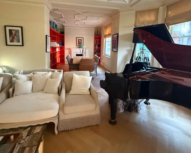 The image shows a well-lit living room featuring a grand piano, a light-colored sofa, and red shelving units along the walls.