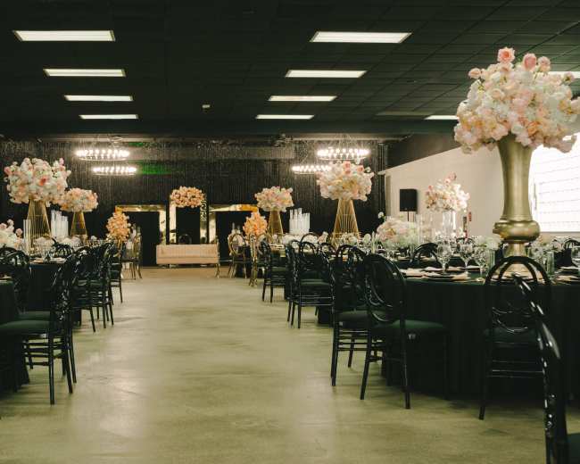 The image shows an elegantly arranged banquet hall featuring black tables set with dinnerware and tall floral centerpieces.