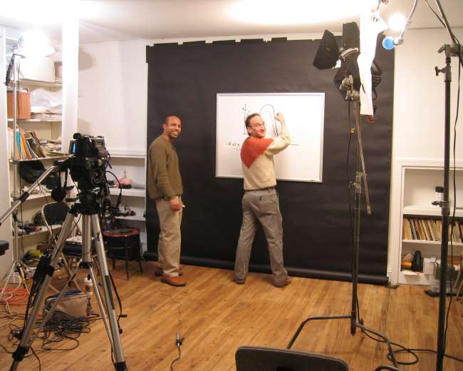 Two men stand in a studio in front of a black backdrop, one drawing on a whiteboard while the other looks on, with cameras and lights set up around them.
