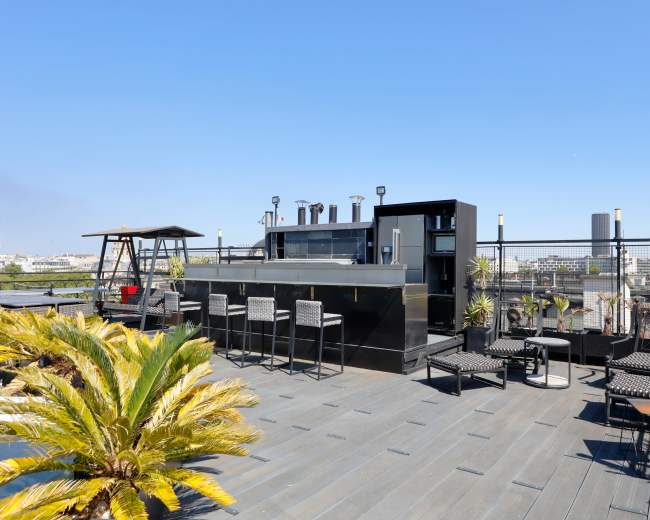 The image shows a rooftop terrace featuring a bar area, seating, and potted plants against a clear blue sky.