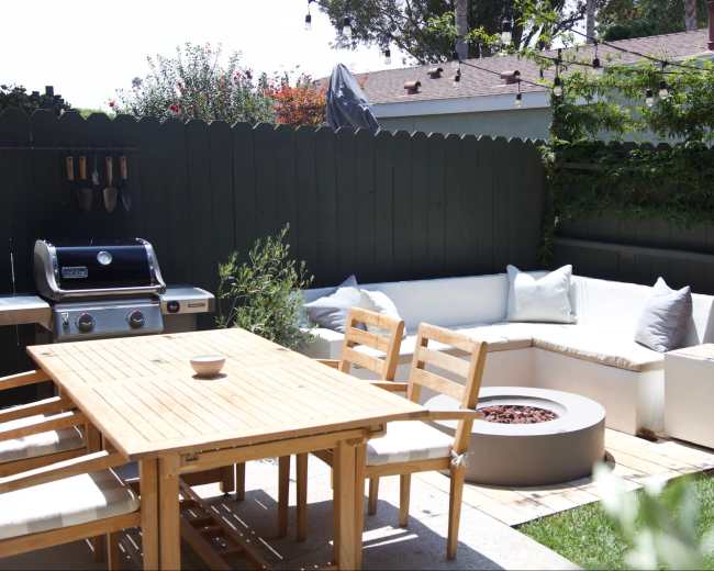 The image shows a backyard patio with a wooden dining table, a sectional sofa, a fire pit, and a grill surrounded by lush greenery.
