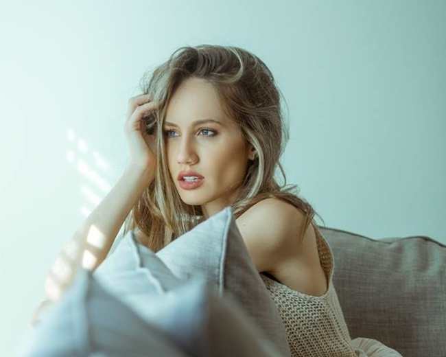 A woman sits on a couch, gazing thoughtfully while resting her hand on her hair, with soft light filtering through nearby blinds.