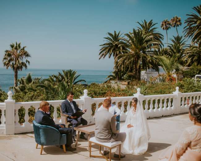 A couple stands in front of a small gathering during a wedding ceremony with a backdrop of palm trees and the ocean.