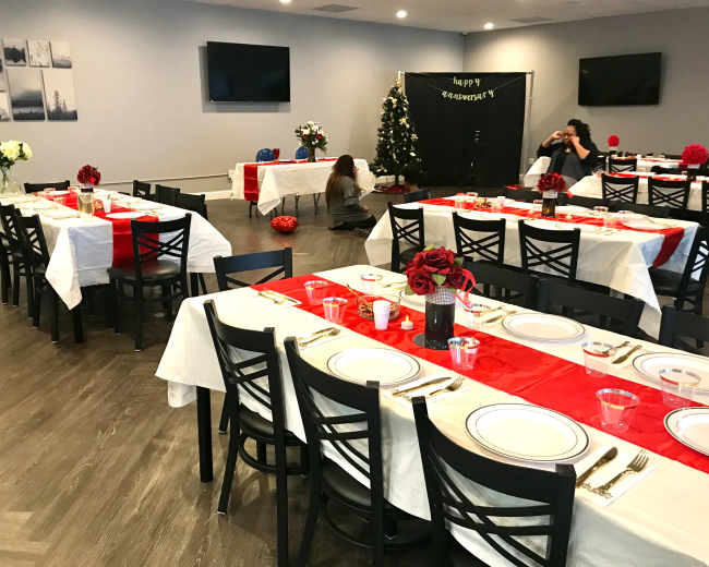 A spacious dining area is set up for a celebration, featuring tables draped in white and red tablecloths, with floral centerpieces and a Christmas tree in the corner.