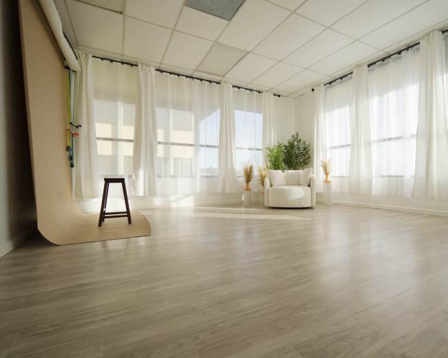 A bright, spacious room with large windows, white curtains, and a white armchair accompanied by potted plants and minimal decor on a wooden floor.