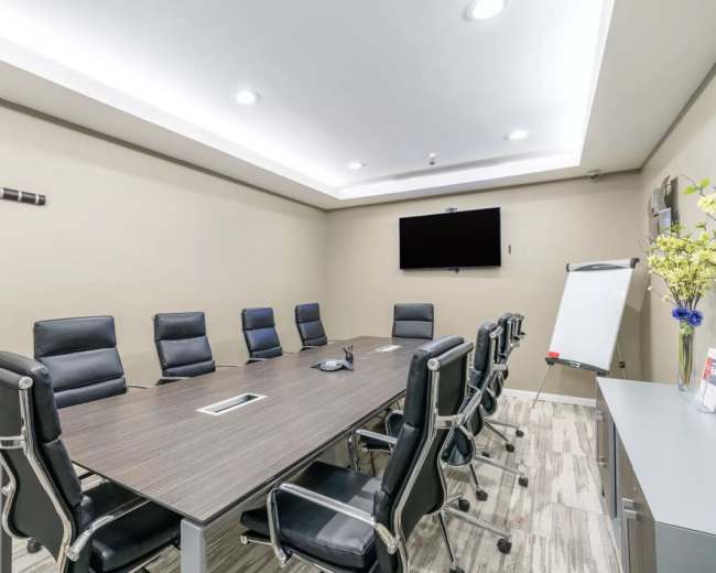A modern conference room features a long table surrounded by black leather chairs, a flat-screen television on the wall, and a whiteboard on a stand.