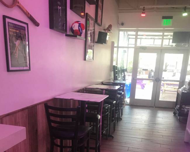 The image shows a narrow interior space with several tall bar stools and tables alongside a wall, leading to a set of double glass doors.