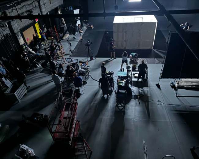 A film crew sets up equipment and prepares for a production in a large, dimly lit studio, with various tools and personnel scattered around.