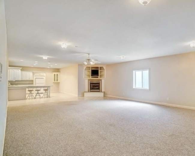 The image shows a spacious living room with light-colored carpeting, a fireplace, and a view into a kitchen area with bar seating.