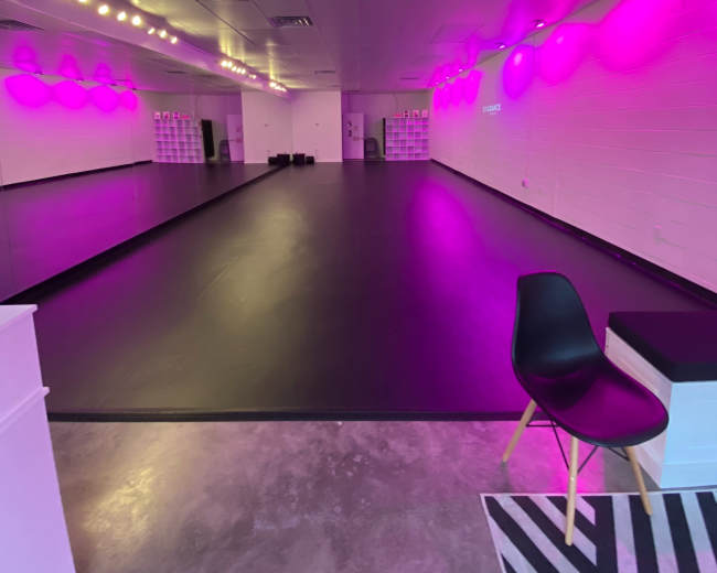 A dance studio with a large black floor, mirrored walls, and pink ambient lighting.