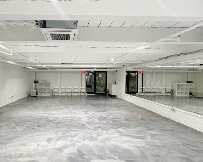 The image shows an empty, well-lit studio space with gray flooring and a large mirror on one wall.