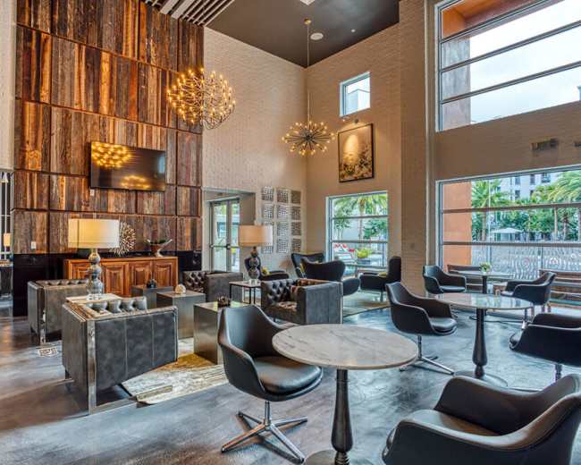 The image shows a modern lounge area featuring stylish black chairs, a wooden wall, a chandelier, and large windows that let in natural light.