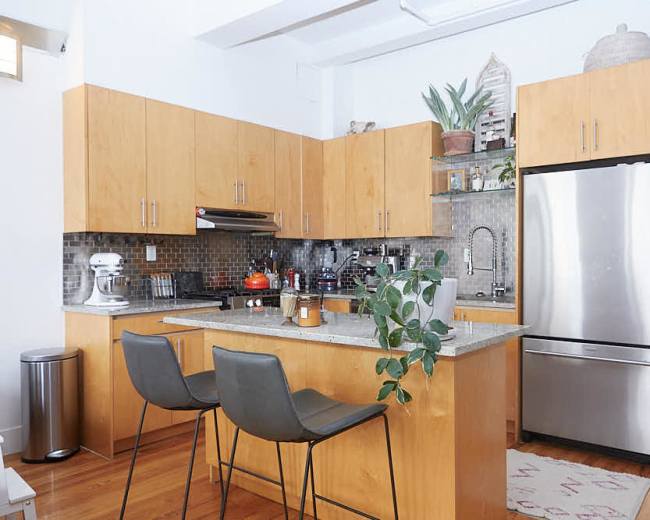 A modern kitchen features light wood cabinetry, a stainless steel refrigerator, and a central island with two stools.