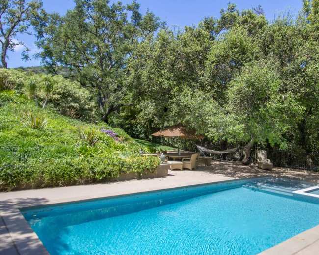 A swimming pool is surrounded by greenery, with a shaded seating area and a hammock nearby.