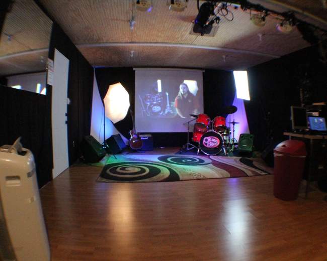 A music rehearsal space with a drum set, guitar, and various lighting setups, along with a computer and a video display.