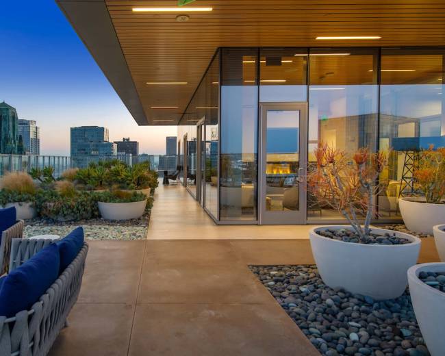 The image shows a modern rooftop terrace with potted plants, lounge seating, and a panoramic view of the city skyline at dusk.