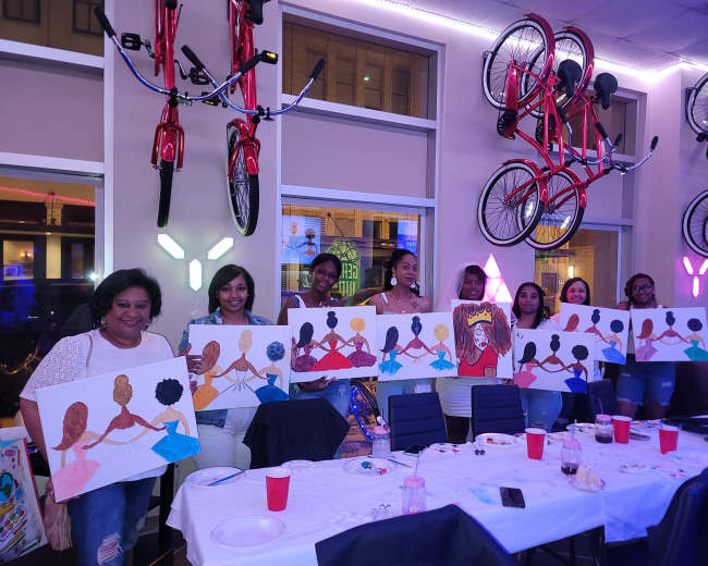 A group of women stands together holding painted canvases, displaying their artwork of figures with varied hairstyles and colors in a decorated venue with bicycles hanging from the ceiling.