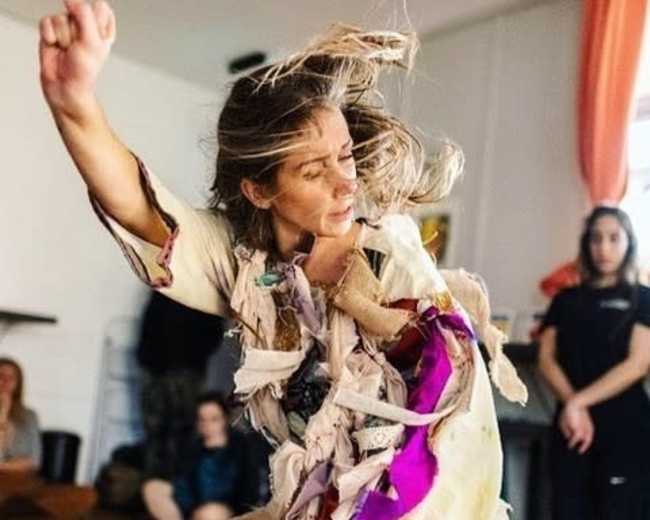 A dancer performs in a studio, wearing a garment made of torn fabric, while onlookers sit on the floor nearby.