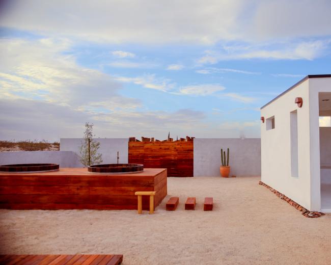 A modern outdoor space featuring wooden hot tubs, a pathway, a potted cactus, and a minimalistic white building against a cloudy sky.