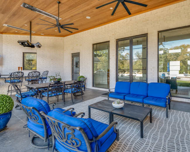The image shows a covered outdoor patio area featuring blue seating, a coffee table, and large glass windows overlooking a garden.