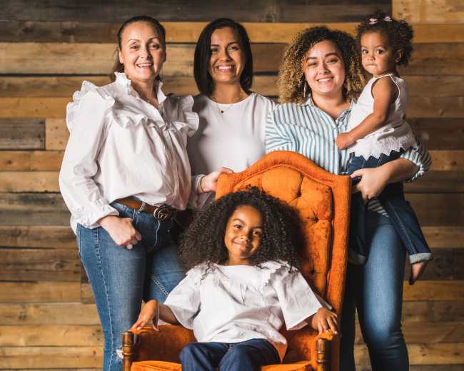 Five women and children pose for a family photo in front of a wooden backdrop, with one girl seated in an orange chair.
