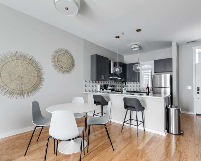 A modern kitchen and dining area features a round table surrounded by chairs, with light-colored cabinetry and decorative wall elements.