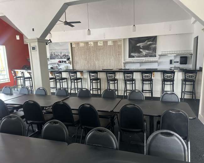 The image shows a dining area with several black tables and chairs, featuring a bar counter in the background with a row of high stools.