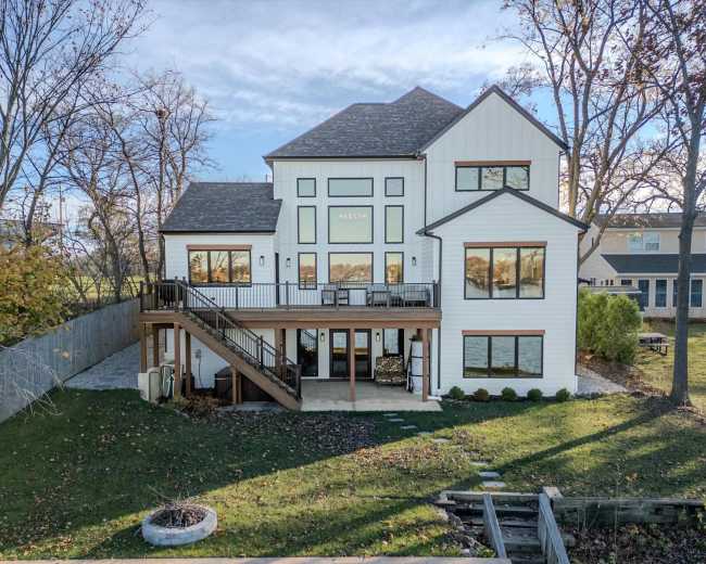 A modern two-story house with large windows and a wooden deck overlooks a lawn bordered by trees.