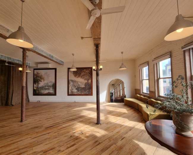 The image shows a spacious, well-lit room with wooden floors, large windows, and two large framed artworks on the walls.