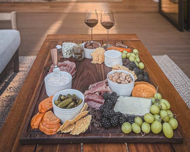 A wooden table is adorned with a charcuterie board featuring assorted meats, cheeses, fruits, and two glasses of red wine, with a cozy living room visible in the background.
