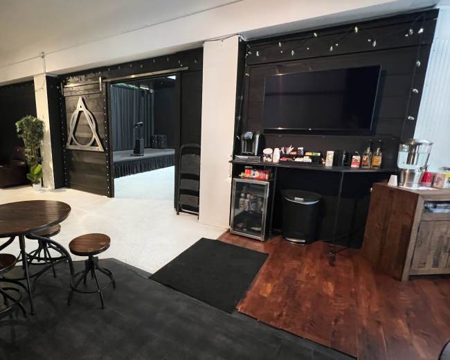 A modern indoor space featuring a seating area with small tables, a kitchenette with a variety of beverages, and an open area leading to a stage-like platform in the background.