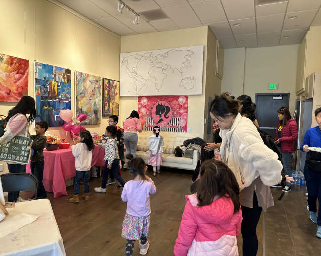 A group of children and adults gather inside a brightly lit room decorated for a party, with pink-themed decorations and artwork on the walls.