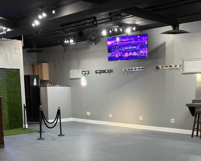 The image shows an empty, gray-floored space with a bar area, a small kitchen to the left, and a television mounted on the wall displaying colorful lights.