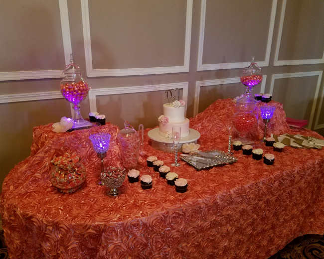 A decorated table covered with a floral-patterned cloth, displaying a tiered cake, various candies in jars, and cupcakes arranged on a silver platter.