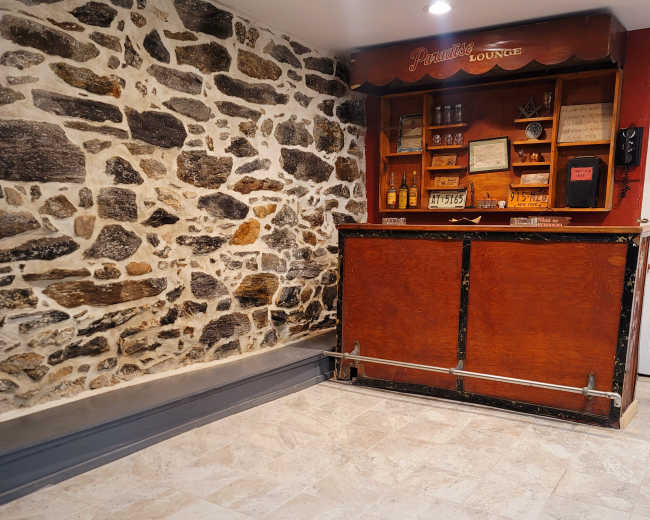 A lounge area with a stone wall, a wooden bar counter, and shelves displaying various bottles and framed items.
