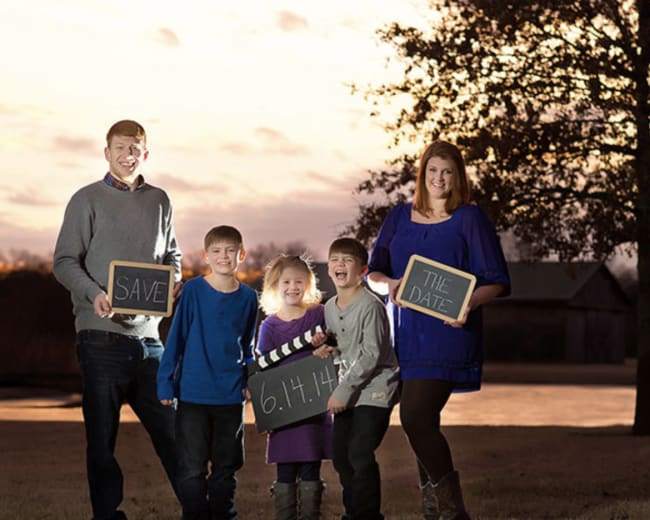 A family stands outdoors holding chalkboards that read "SAVE," "THE DATE," and "6.14.14" against a backdrop of a sunset.