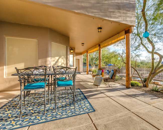 A patio area with a table and chairs, bordered by desert landscaping and shaded by a building.