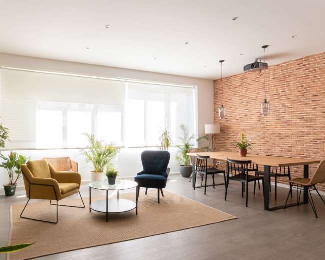 The image shows a modern living space featuring a beige and brick wall, a round table with chairs, and various plants arranged around the room.