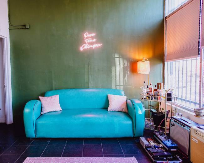 A teal sofa is positioned against a green wall with a neon sign, while nearby furniture and decor create a cozy living space.