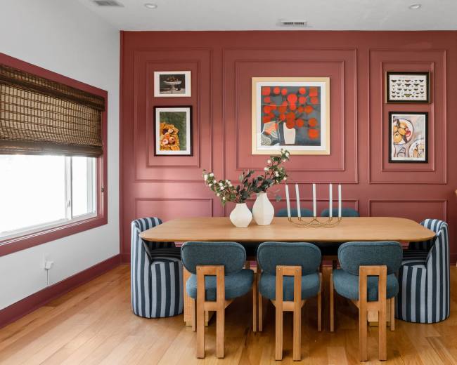 The image shows a dining area with a rectangular wooden table surrounded by chairs, a red accent wall featuring framed artwork, and a kitchen area visible in the background.