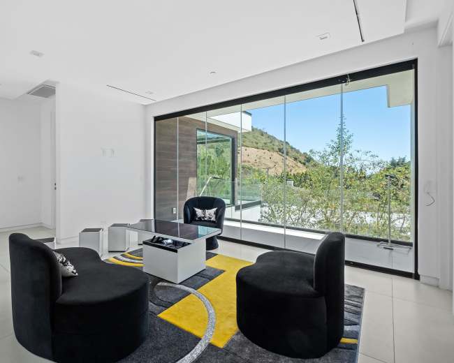 A modern living room features two black chairs and a glass table, with large windows overlooking a hillside landscape.