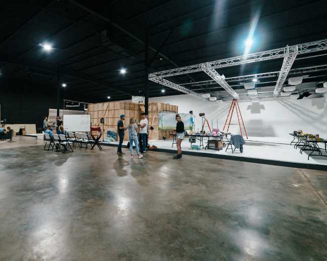 A group of people are engaged in various activities within a spacious studio featuring a polished concrete floor and backdrop sets in the background.