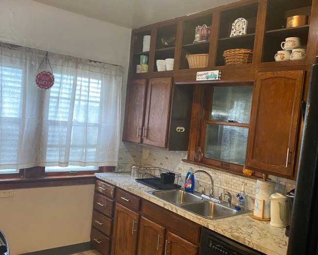 The image shows a kitchen with wooden cabinets, a sink, and a window adorned with sheer curtains.