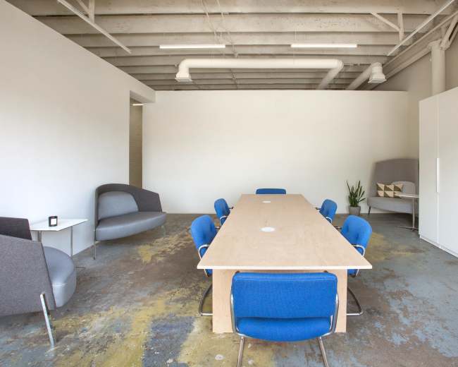A long wooden table surrounded by blue chairs is positioned in a spacious, minimalistic meeting room with gray furniture and concrete flooring.