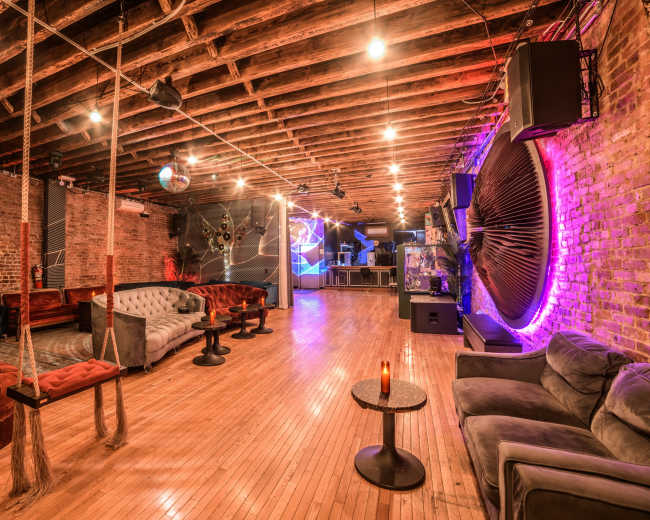 The image shows a spacious, brightly lit lounge with wooden floors, exposed beams, and a mix of seating options including sofas and chairs.