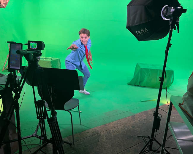 A person in a blue outfit and red tie poses playfully in front of a green screen, surrounded by filming equipment.