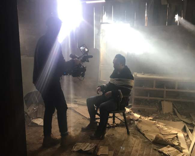 A man is tied to a chair in a dimly lit room while a person uses a camera to film him.
